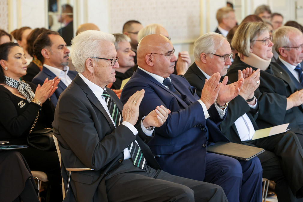 Ministerpräsident Winfried Kretschmann (vorne links) und Daniel Strauß (vorne, zweiter von links), Landesvorsitzender des Verbands Deutscher Sinti und Roma, sitzen im Publikum und applaudieren.